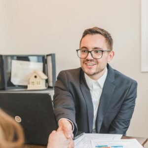 A professional real estate agent welcomes a client in the office with documents on the desk.